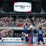 2026年天皇杯・皇后杯全日本バレーボール選手権大会の試合会場の様子。ネット際での迫力あるスパイクシーンと、日本国旗を掲げて応援する満員の観客席。