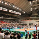 2026年 天皇杯・皇后杯 全日本バレーボール選手権大会の試合会場の様子。満員の観客が詰めかけたアリーナで熱戦を繰り広げるバレーボール選手と、日の丸を振って応援するファンの風景。