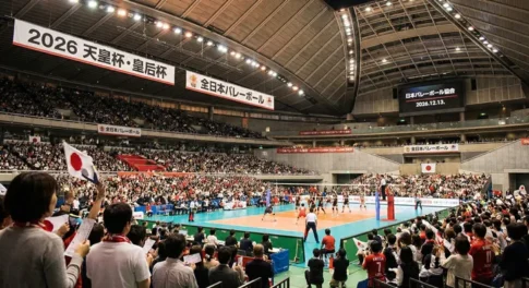 2026年 天皇杯・皇后杯 全日本バレーボール選手権大会の試合会場の様子。満員の観客が詰めかけたアリーナで熱戦を繰り広げるバレーボール選手と、日の丸を振って応援するファンの風景。