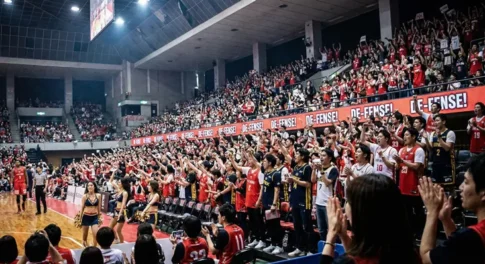 バスケットボールの試合会場で「DE-FENSE!（ディフェンス）」のサインを掲げて熱狂的に応援する観客と、サイドラインで踊るチアリーダーの様子