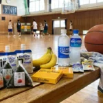 バスケットボールの試合日のエネルギー補給食。体育館のベンチに置かれたおにぎり、バナナ、エネルギーゼリー、カロリーメイト、ポカリスエット、アクエリアスとバスケットボール。