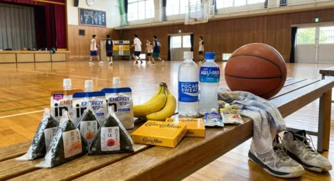 バスケットボールの試合日のエネルギー補給食。体育館のベンチに置かれたおにぎり、バナナ、エネルギーゼリー、カロリーメイト、ポカリスエット、アクエリアスとバスケットボール。