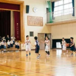 小学校の体育館でミニバスケットボールの試合をする子供たち。一般のバスケとのルールの違いやコート、リングの高さの違いを解説するイメージ。
