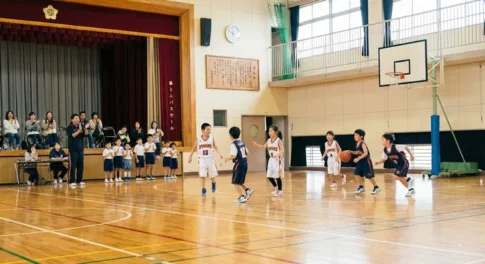 小学校の体育館でミニバスケットボールの試合をする子供たち。一般のバスケとのルールの違いやコート、リングの高さの違いを解説するイメージ。