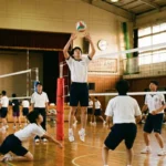 体育館でバレーボールの練習試合をする男子高校生たち。セッターがジャンプトスを上げ、ネット際のブロッカー2人がジャンプして守備につき、レシーバーが構えているプレー風景