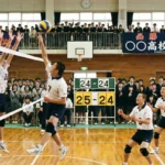 高校バレーボールの試合で24-24のデュースから25-24へリードした緊迫の場面。ネット際でのスパイクとブロックの攻防。25点先取や2点差ルール、セットポイントを解説する記事に適した、観客が見守る白熱した試合風景