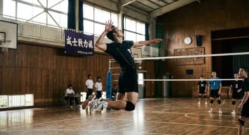 バレーボールのジャンプサーブを打つ瞬間の男子選手。体育館のコートで高くジャンプし、ネット越しにレシーブを構える相手チームに向かってサーブを放つダイナミックなフォーム。