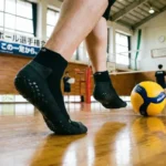 体育館で滑り止め付きのバレーボールソックスを履いて踏ん張る選手の足元。床には公式球が転がり、背景には全日本バレーボール選手権大会の横断幕がある本格的な練習風景。