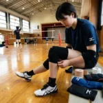 「体育館のベンチでバレーボール用の膝サポーターを装着する女子選手。横にはテーピングや各種サポーターが置かれている」