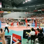 2026年天皇杯・皇后杯バレーボール選手権大会の試合会場。手前でスマホやタブレットを使いライブ配信を視聴する観客と、テレビ中継用のプロ機材、そしてコート上でアタックを放つ選手たちが映る臨場感あふれる風景。