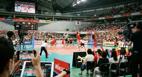 2026年天皇杯・皇后杯バレーボール選手権大会の試合会場。手前でスマホやタブレットを使いライブ配信を視聴する観客と、テレビ中継用のプロ機材、そしてコート上でアタックを放つ選手たちが映る臨場感あふれる風景。