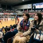 「アリーナの観客席で、双眼鏡や一眼レフカメラを手にVリーグのバレーボール試合を笑顔で観戦する2人の女性ファン」