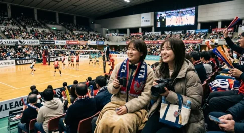 「アリーナの観客席で、双眼鏡や一眼レフカメラを手にVリーグのバレーボール試合を笑顔で観戦する2人の女性ファン」